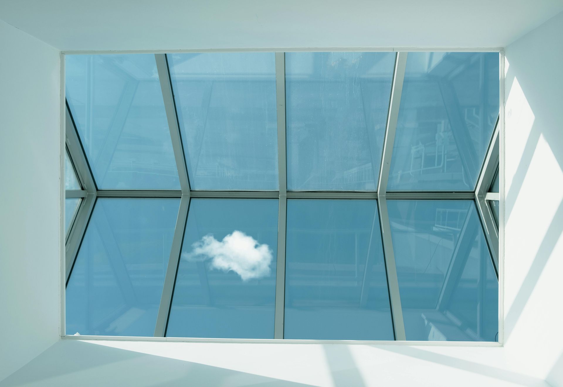 Looking up through a large skylight at a blue sky with a cloud. White frame inside a white room.
